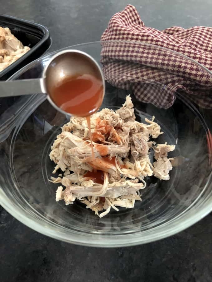 Adding hot wing sauce to shredded chicken in glass bowl.
