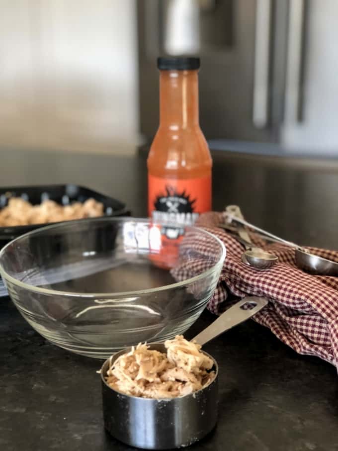 Shredded chicken, glass bowl, hot wing sauce and measuring spoons on kitchen counter.