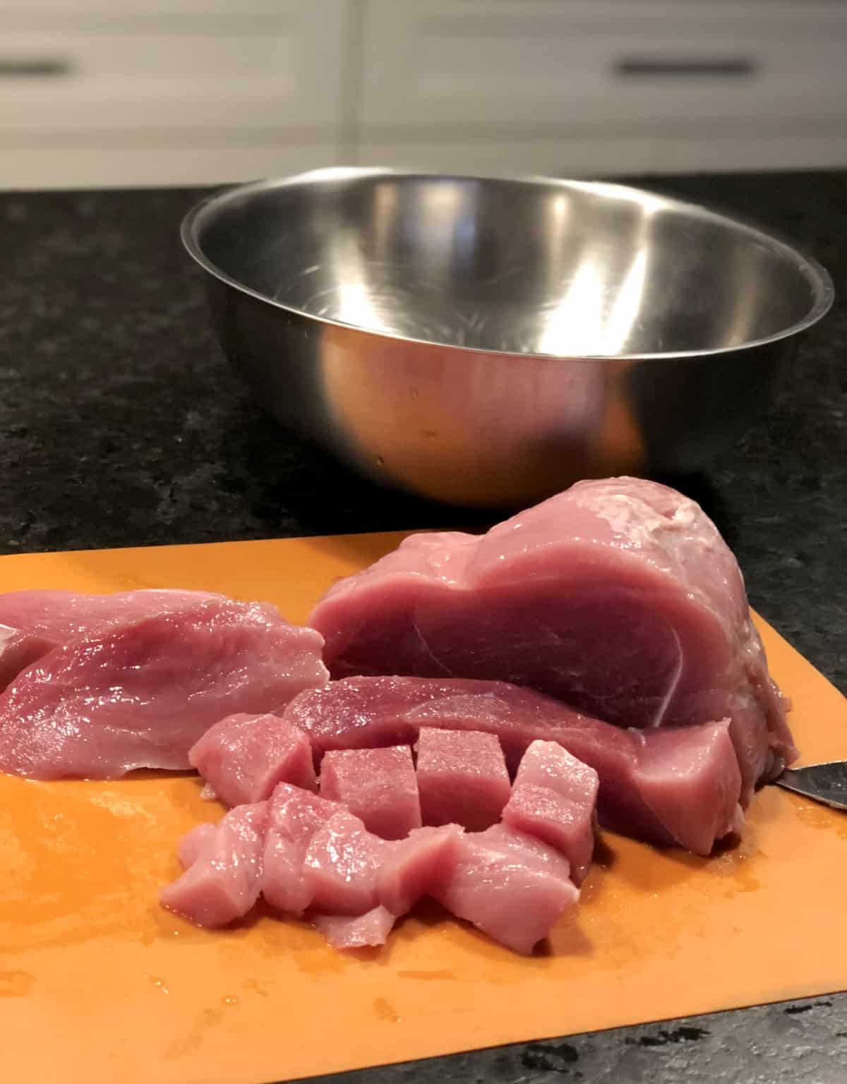 Cutting raw pork roast into cubes on orange cutting mat with stainless bowl in background
