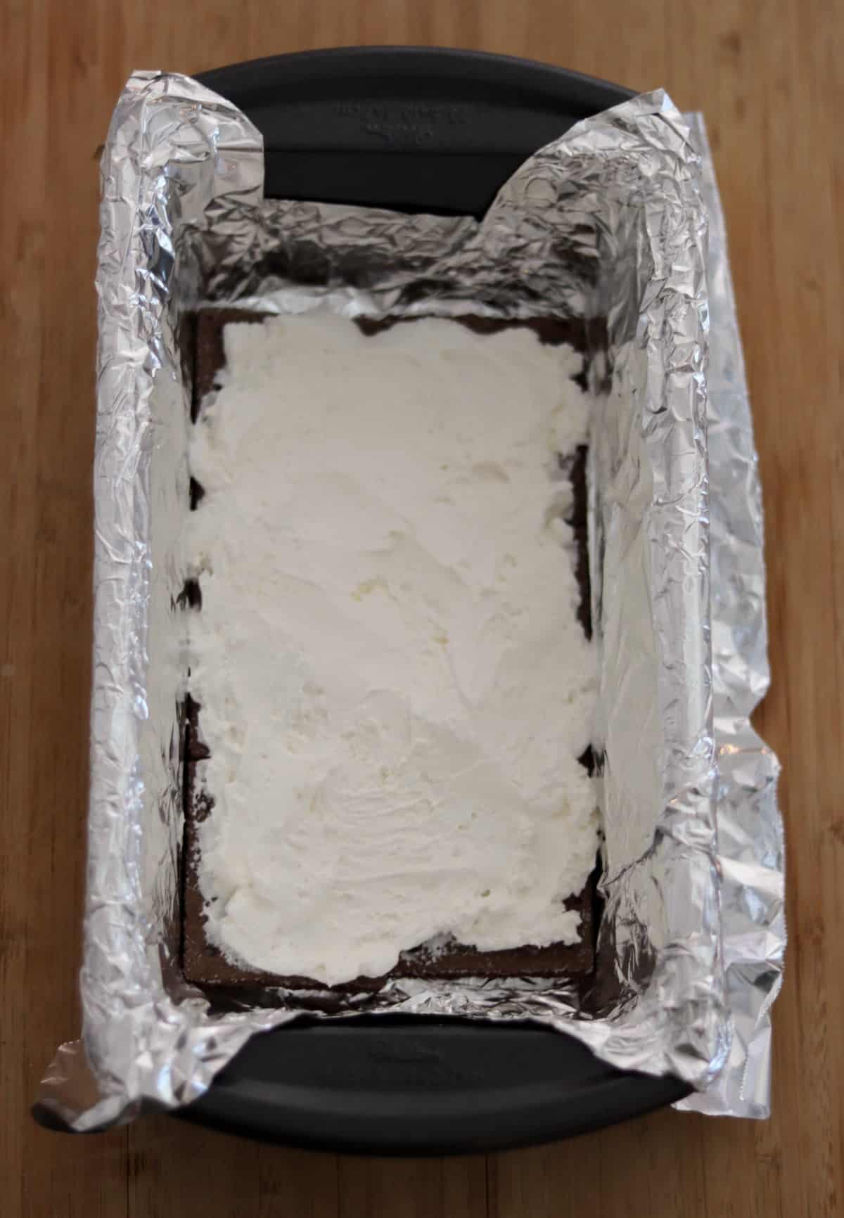 Foil-lined loaf pan with layer of chocolate graham crackers topped with frozen whipped topping.