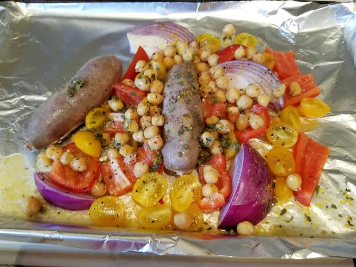 Sausages with corn, red peppers and red onion on foil-lined baking pan.
