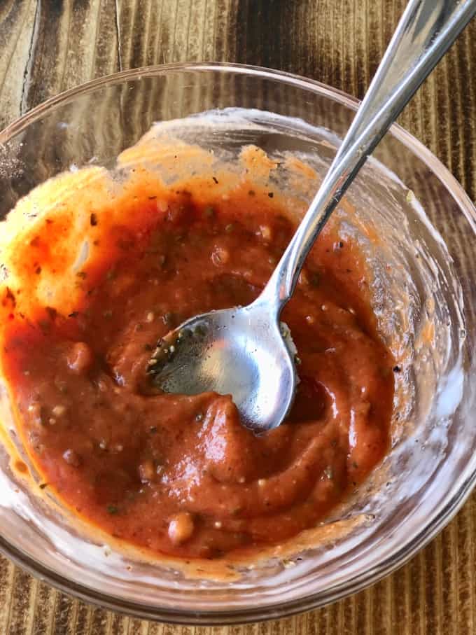 Tomato paste mixed with Italian herbs and cream cheese in small glass bowl with spoon.