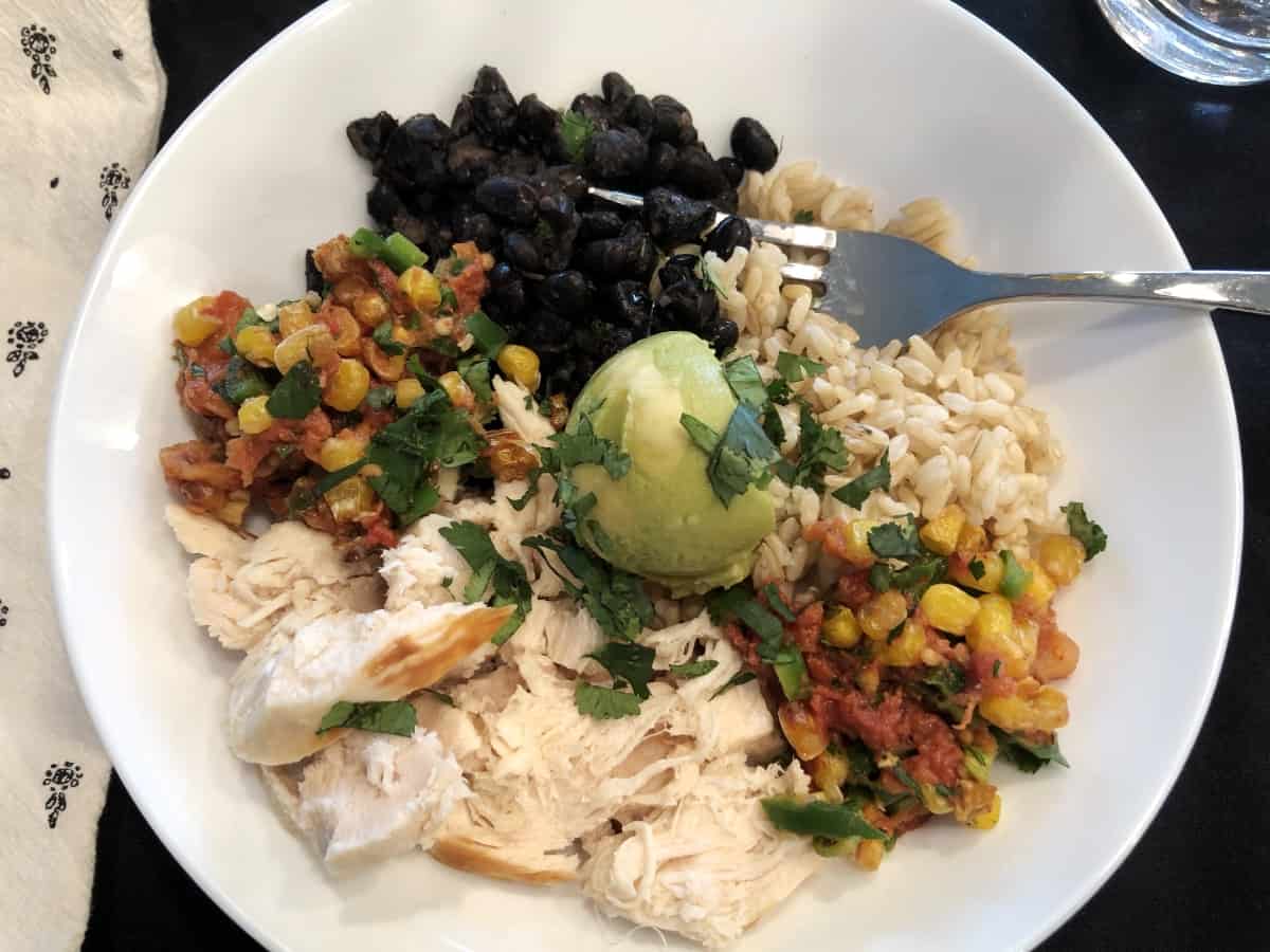 Black beans, brown rice, chopped chicken, corn salsa, avocado and chopped cilantro in bowl with fork.