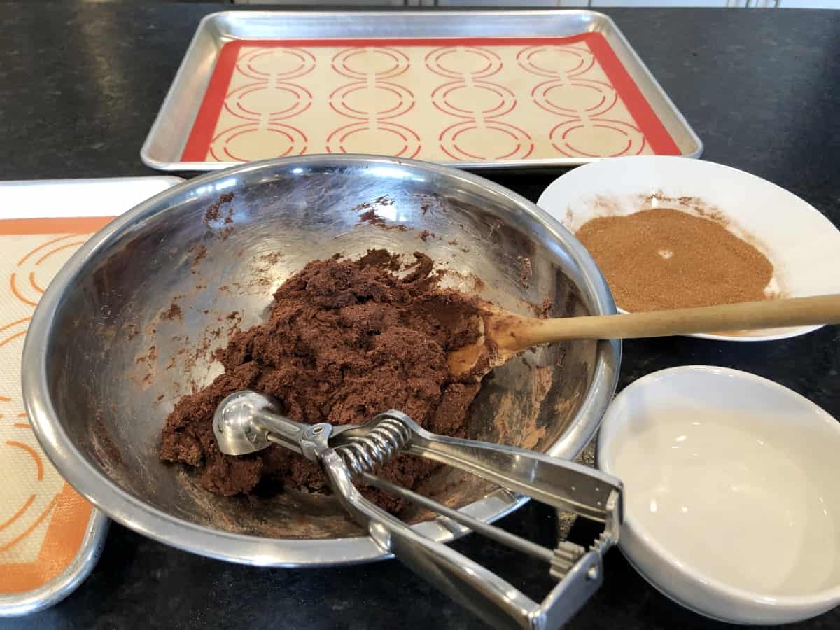 Chocolate cake mix cookie dough in bowl with cookie scoop and wooden spoon and baking sheet in background.