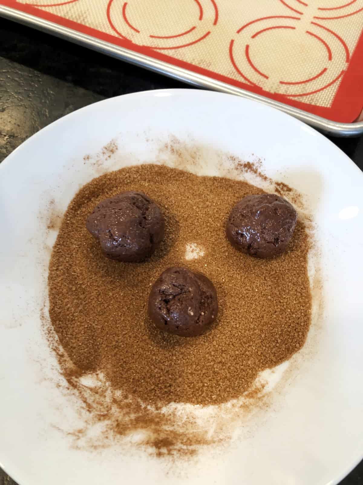 Three chocolate cookie dough balls in cinnamon sugar mixture in shallow white bowl.