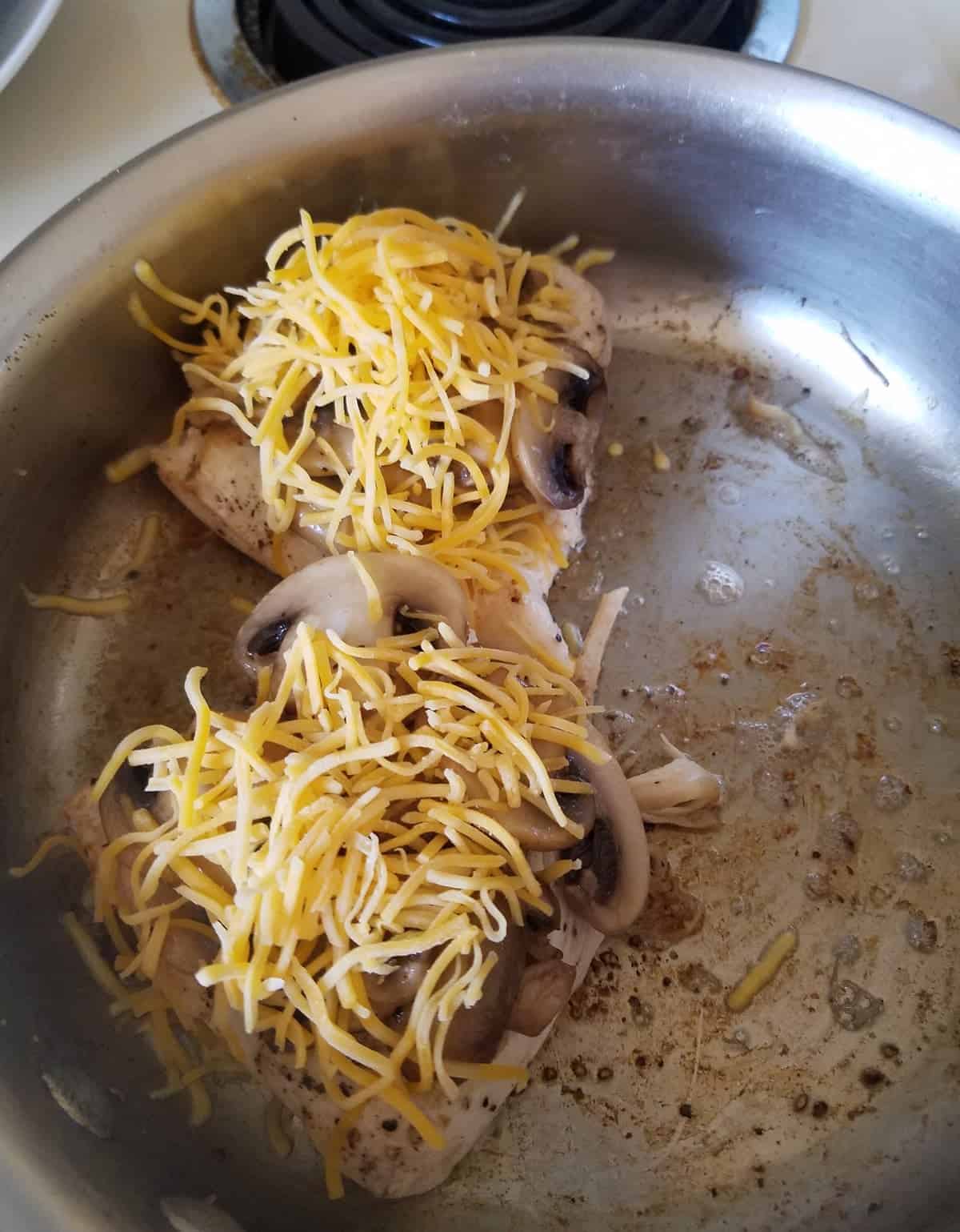 Chicken breasts topped with mushrooms and shredded cheese in saute pan.