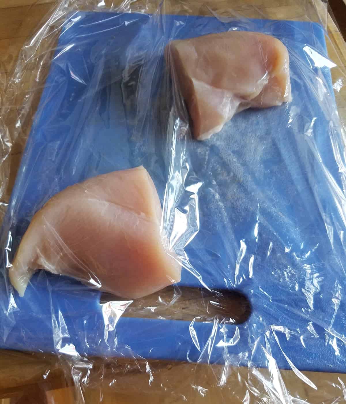 Chicken breasts covered with plastic wrap on blue cutting board