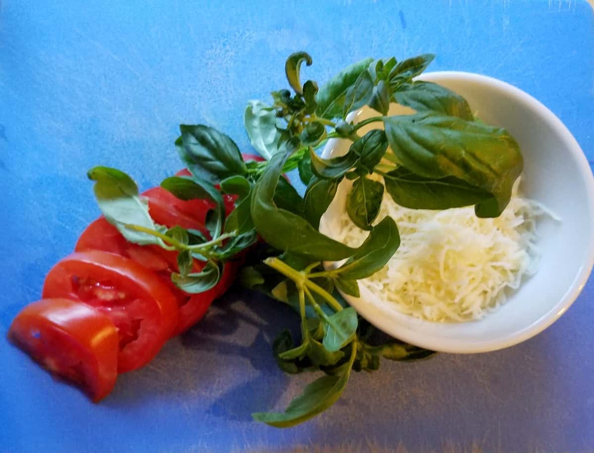 Sliced tomatoes, bunch of basil leaves and shredded mozzarella on blue cutting mat.