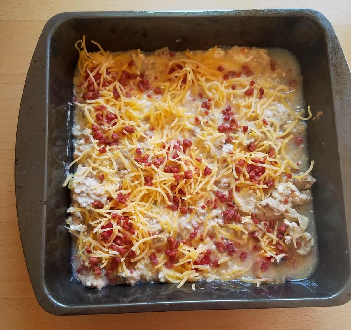 Unbaked cheeseburger casserole topped with shredded cheese and bacon bits in baking dish.