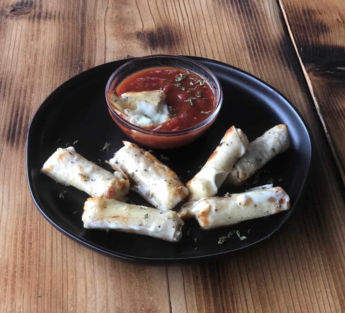 Wonton wrapped mozzarella cheese sticks on black plate with small bowl of marinara dipping sauce on wood table