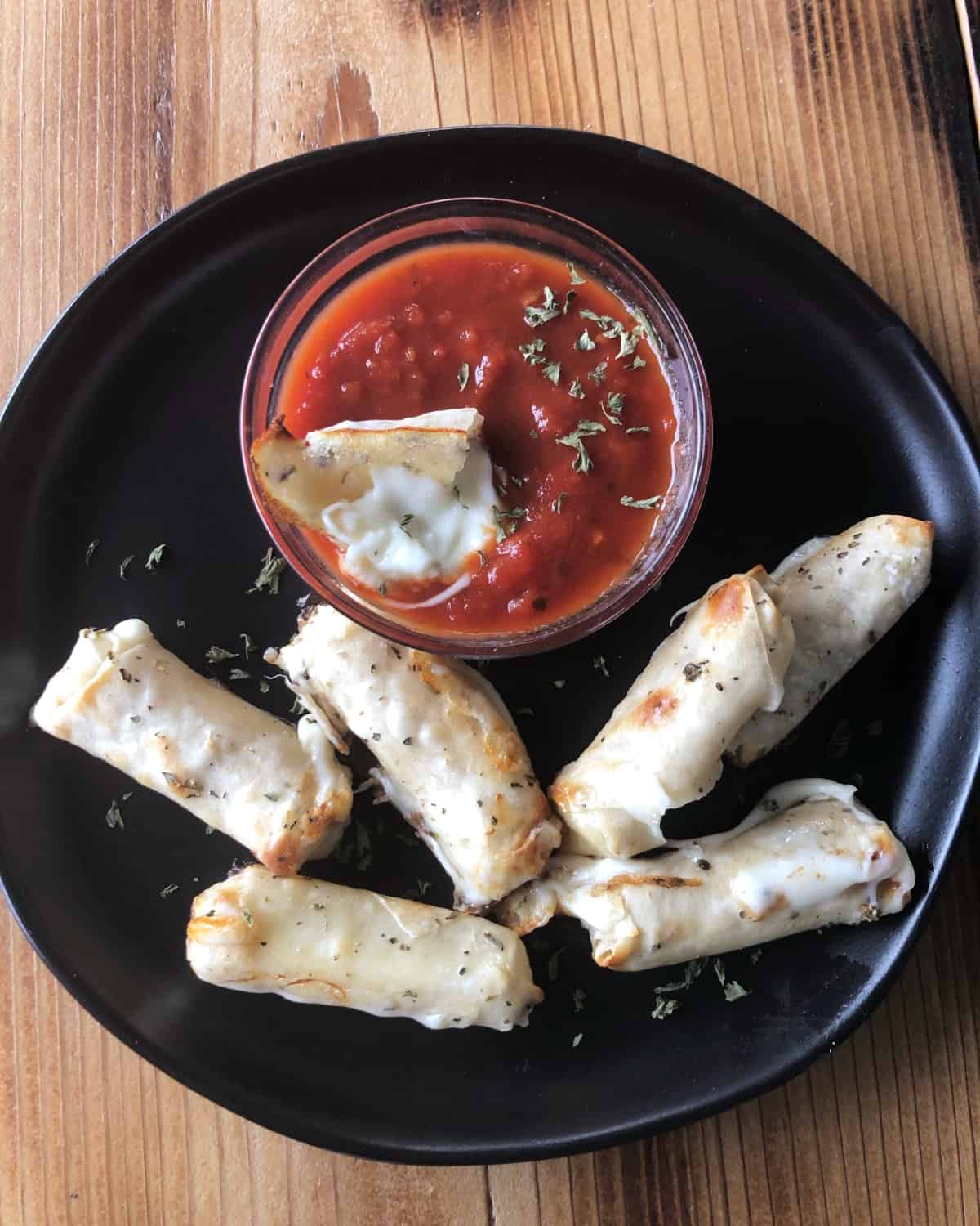 Wonton-Wrapped Baked Cheese Sticks with Marinara Dipping Sauce on black plate.