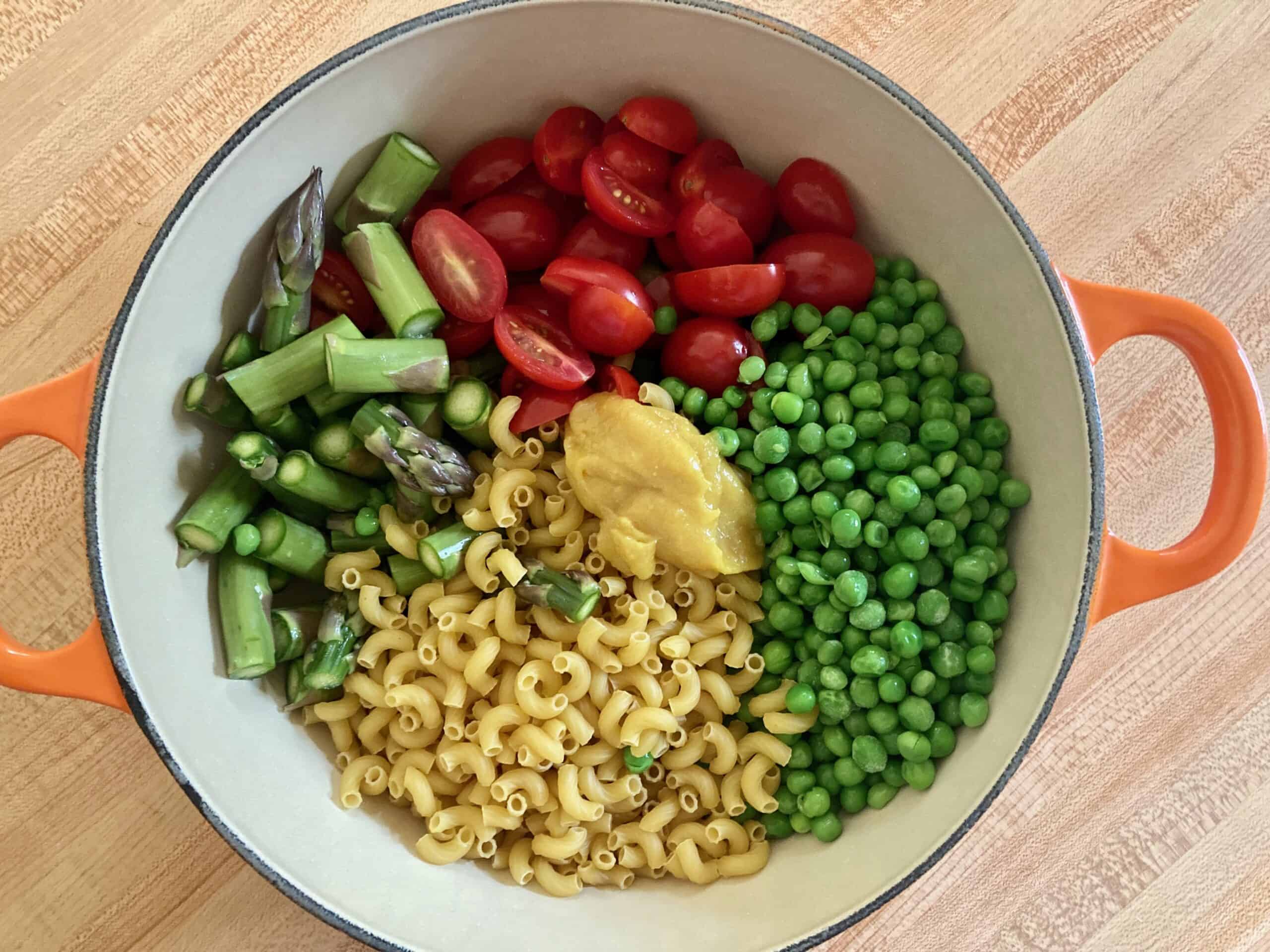 Chopped asparagus, sliced tomatoes, elbow macaroni, peas, and better than bouillon paste in an enameled cast iron soup pot shot from above.