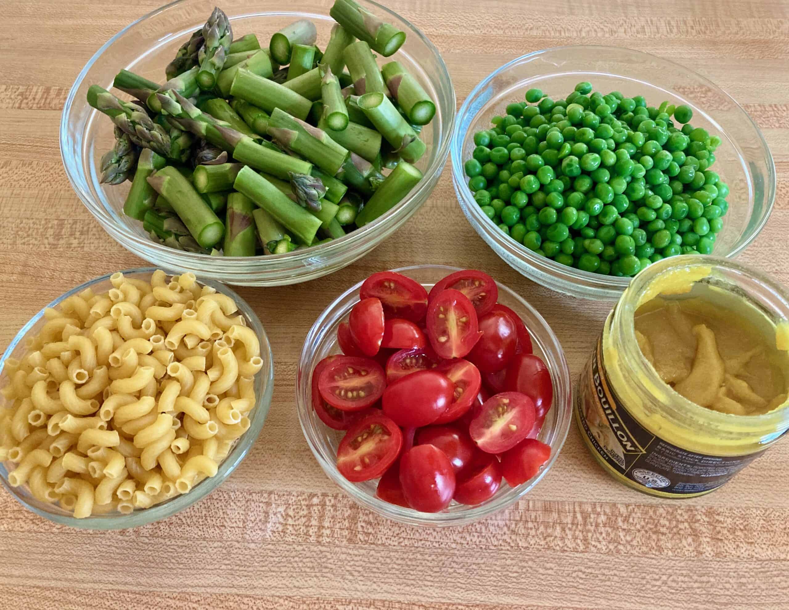 Bowls of chopped asparagus, peas, elbow macaroni and sliced cherry tomatoes on a counter along with a jar of Better Than Bouillon.