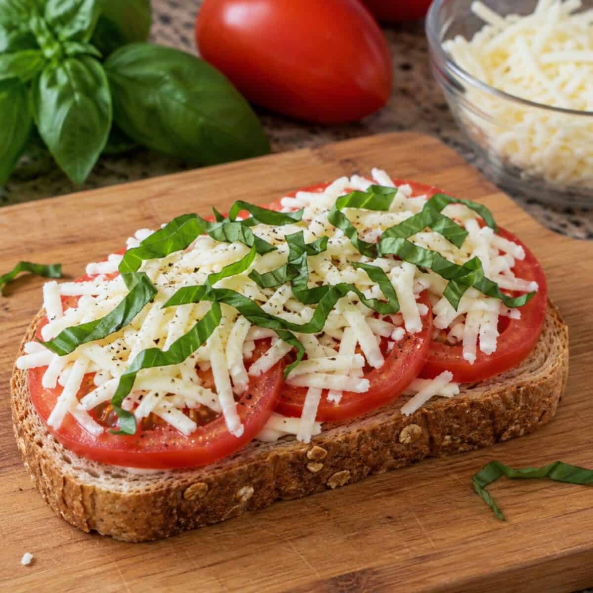 Unbaked open-faced caprese sandwich on whole wheat bread on wooden cutting board.
