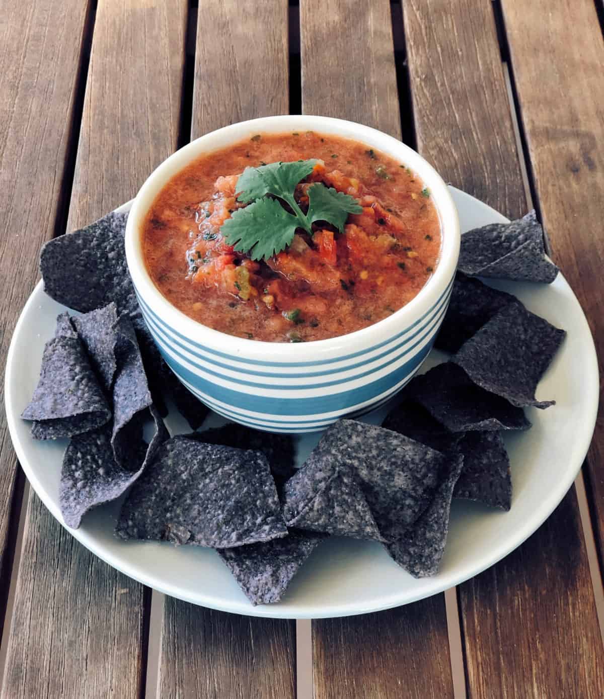 Bowl of fresh homemade salsa on plate with blue corn tortilla chips on wooden table.