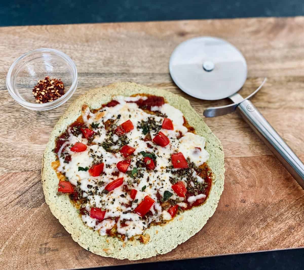 Tortilla pizza on cutting board with pizza cutter and crushed red pepper flakes.