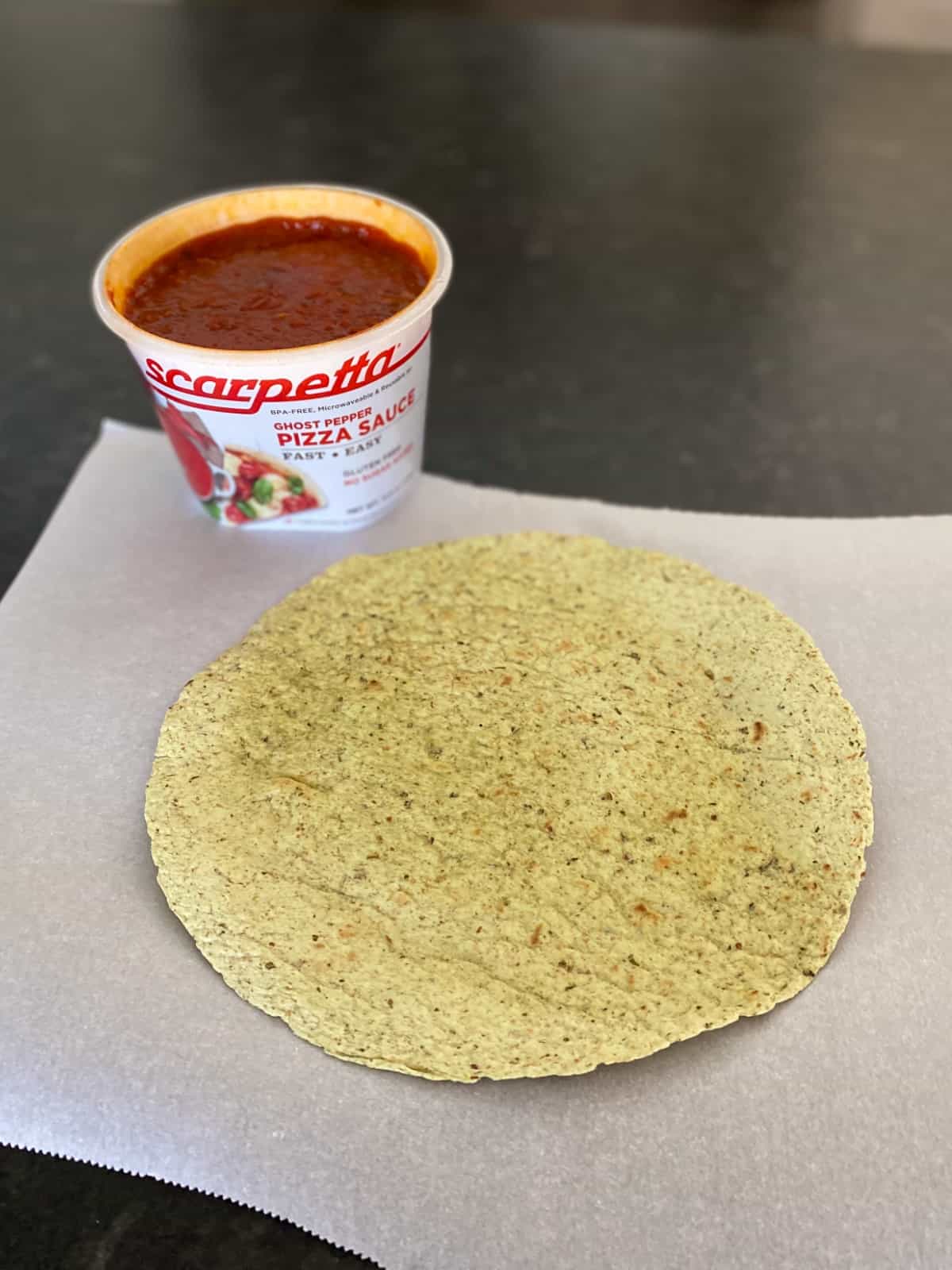 Pizza sauce container and crispy baked tortilla sitting on parchment paper.