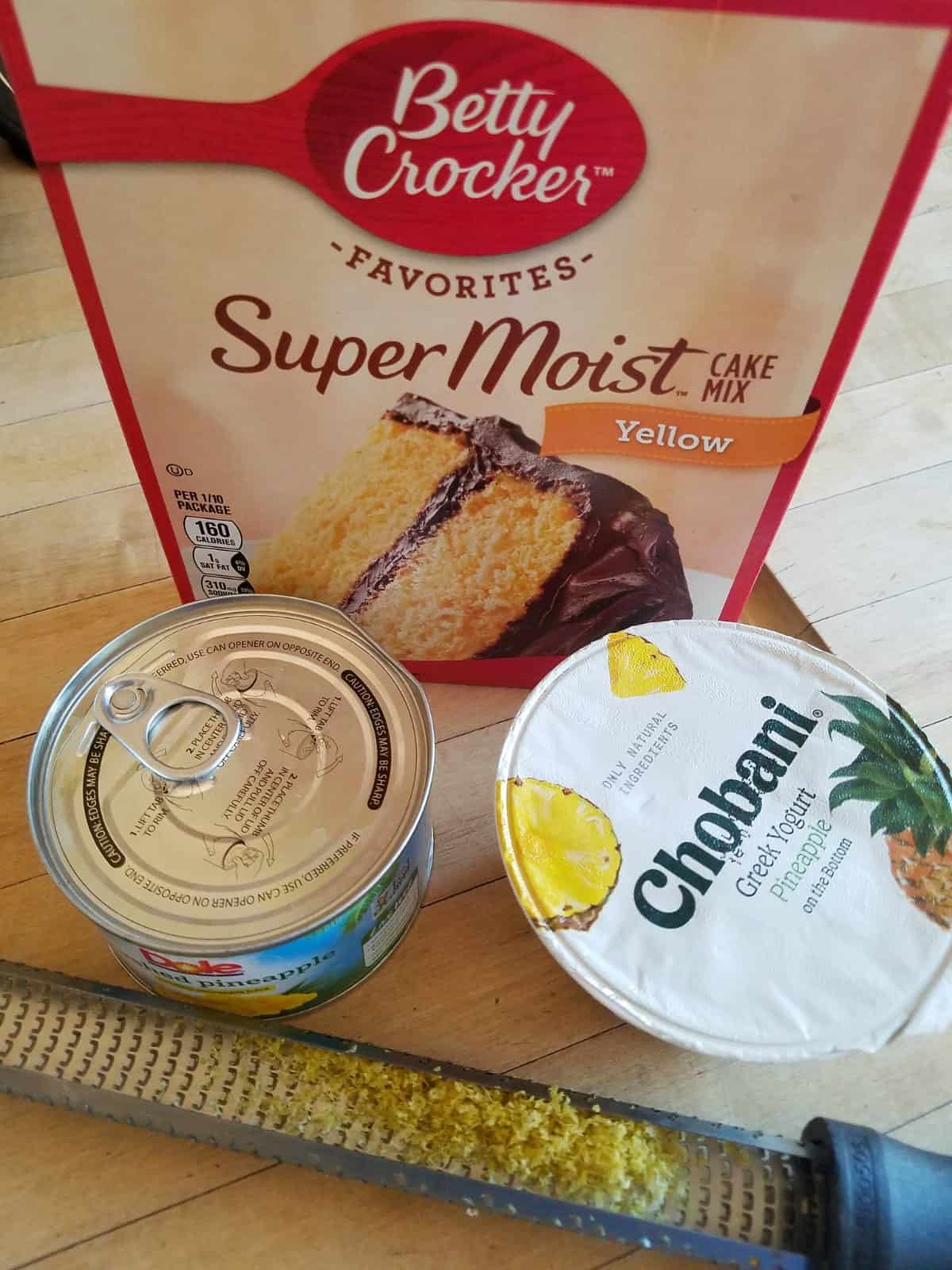 Box of supermoist yellow cake mix, can of crushed pineapple, container of pineapple greek yogurt and lemon zester on counter.