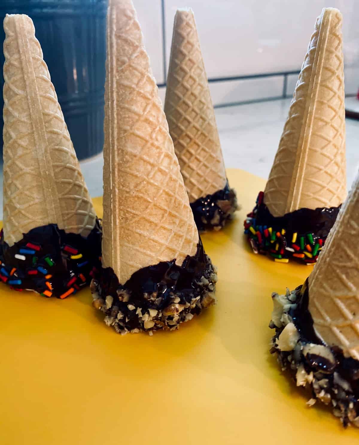 Chocolate dipped ice cream cones with sprinkles and chopped nuts sitting on yellow mat on counter.