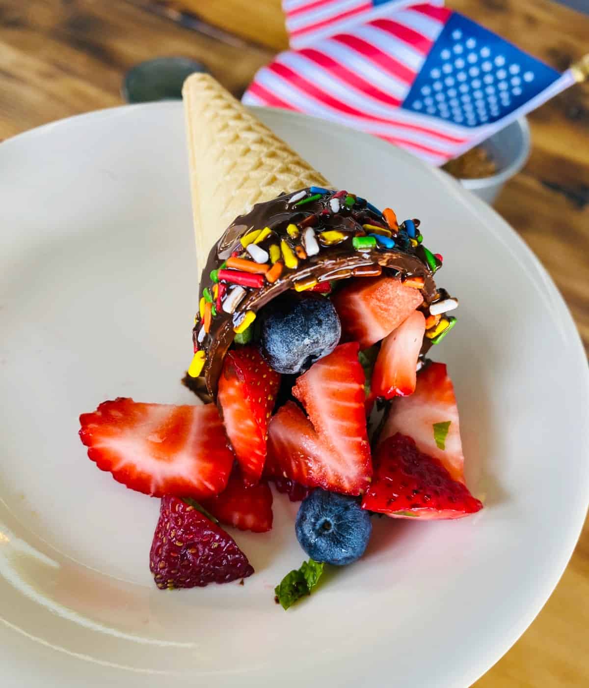 Chocolate dipped ice cream cone filled with fresh berries on white plate with American flag in background.