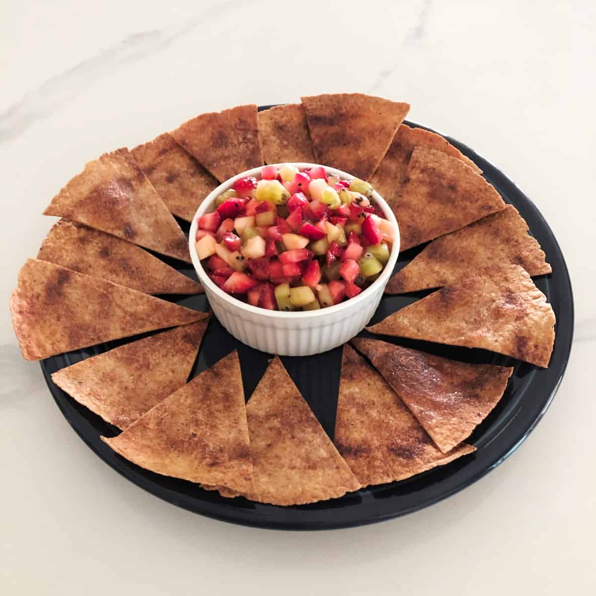 Fruit salsa in white bowl on platter scatter with cinnamon-sugar crisps.