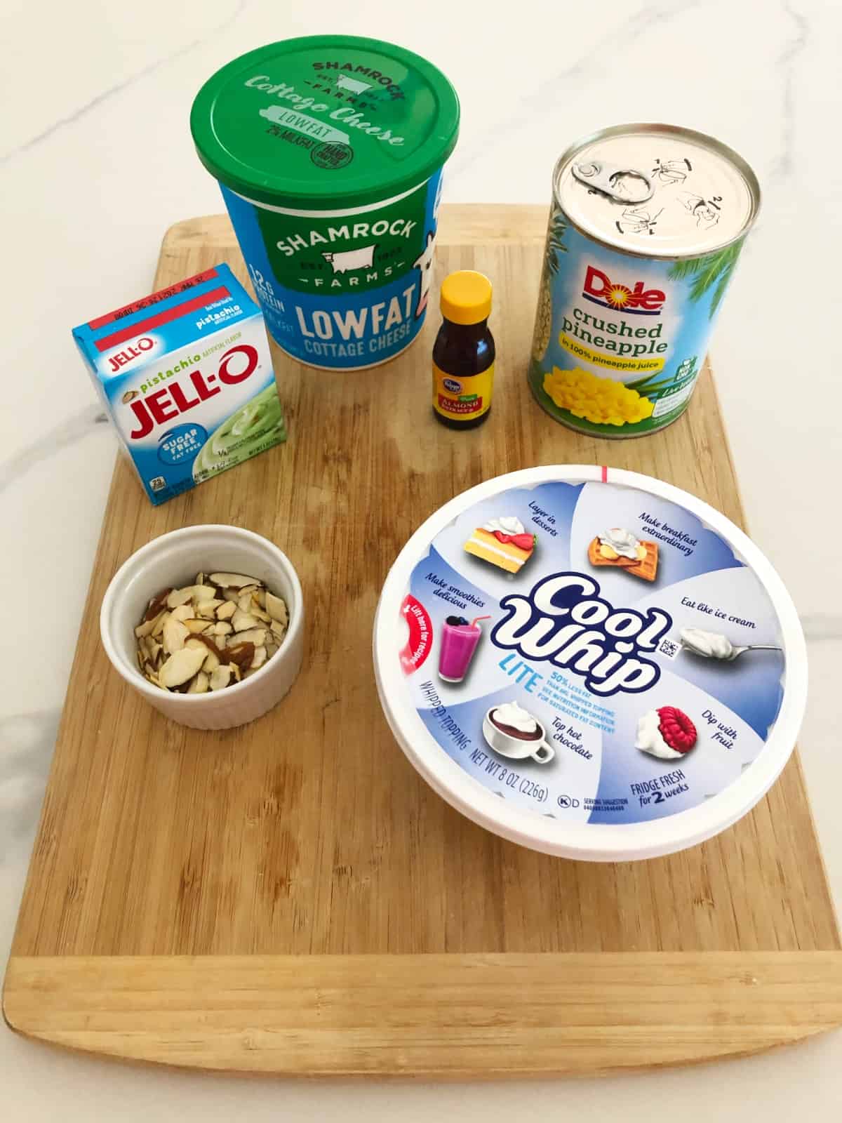 Ingredients on bamboo cutting board including pistachio pudding mix, cottage cheese, pineapple, cool whip and sliced almonds.