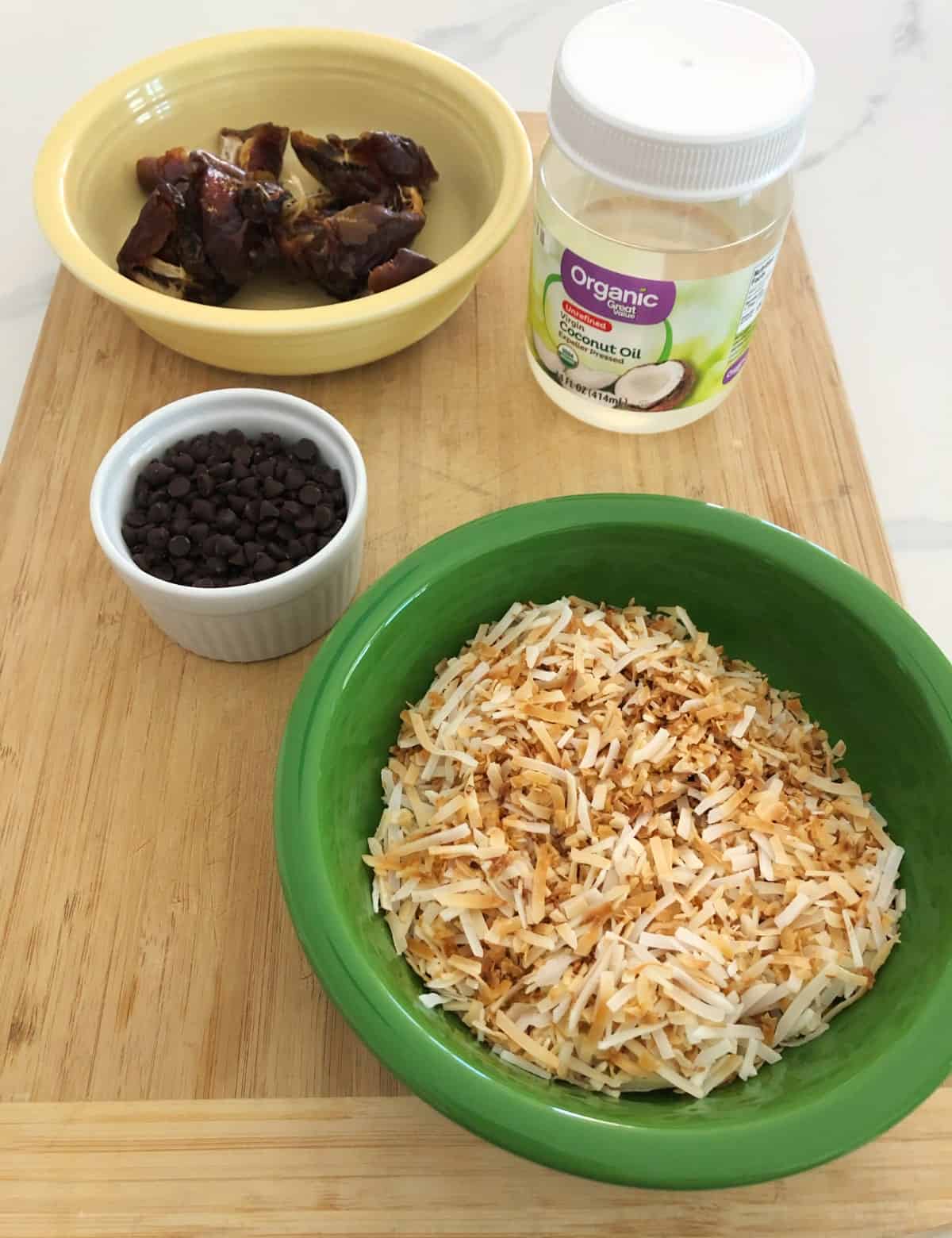Pitted dates, mini chocolate chips, toasted coconut and coconut oil on bamboo cutting board.