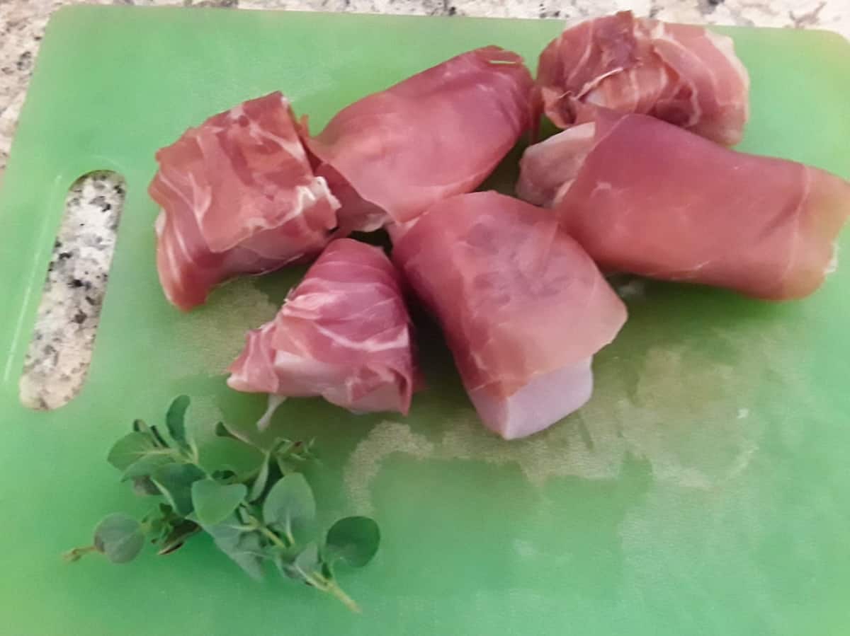 Six pieces chicken breast wrapped in prosciutto on green cutting board with fresh oregano.