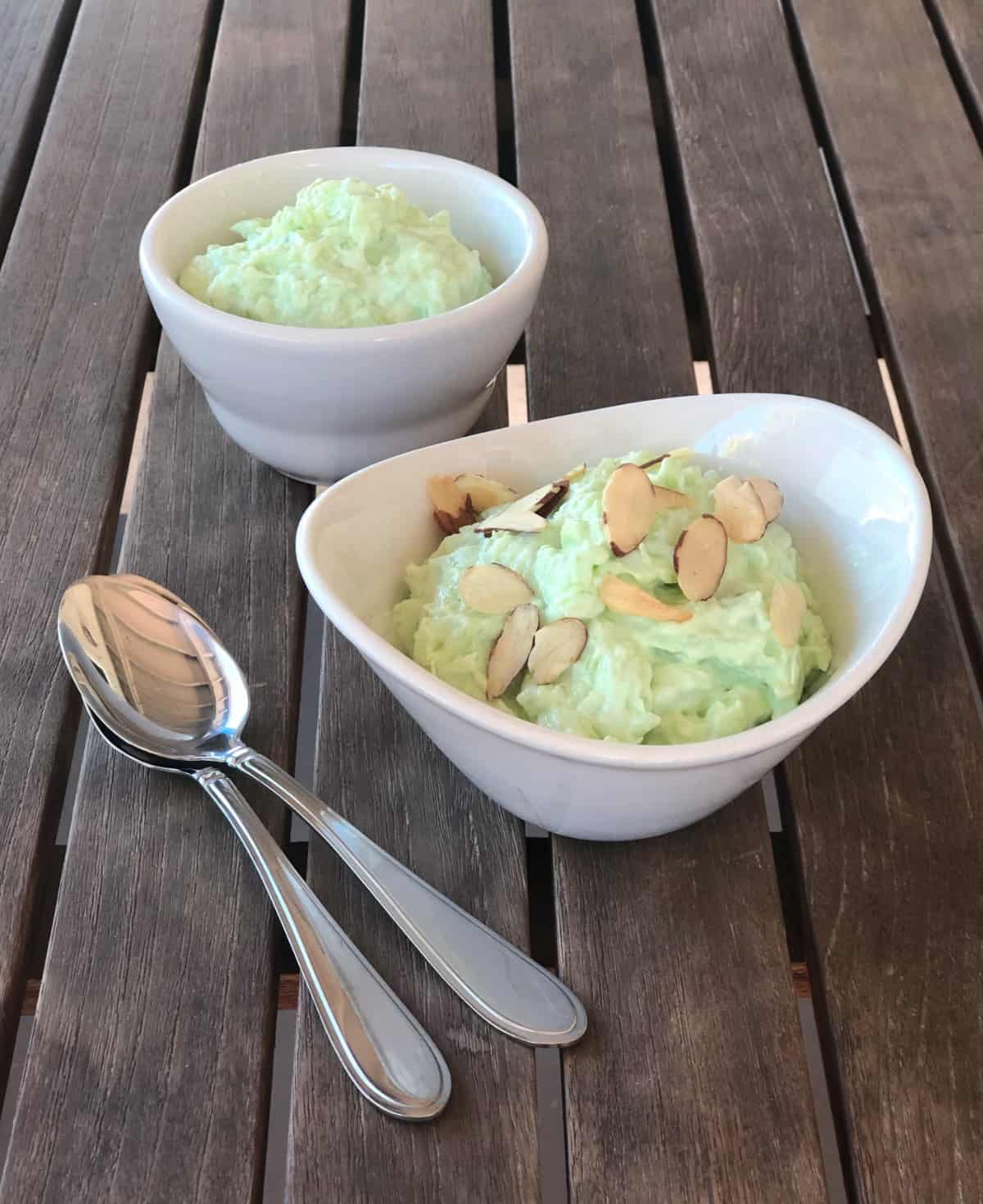 Two small white bowls with Watergate salad, one topped with sliced almonds one without, next to two spoons.