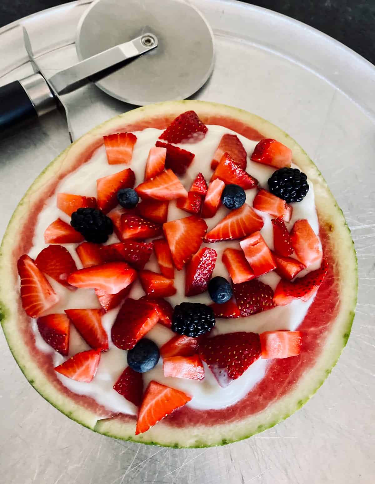 Watermelon pizza topped with yogurt, strawberries, blueberries and blackberries with pizza cutter.