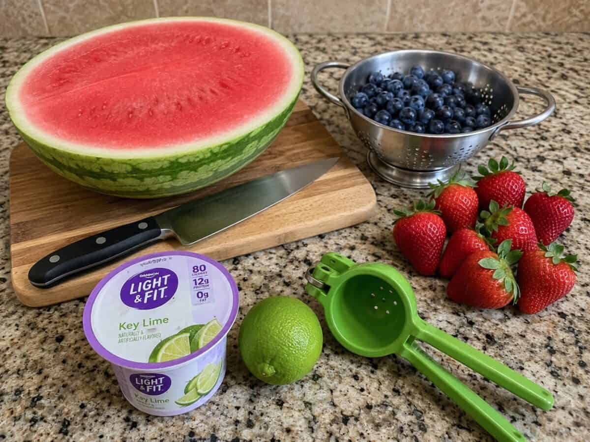 Half a seedless watermelon, blueberries in a colander, fresh strawberries, small container of key lime yogurt, lime and lime squeezer on granite counter.