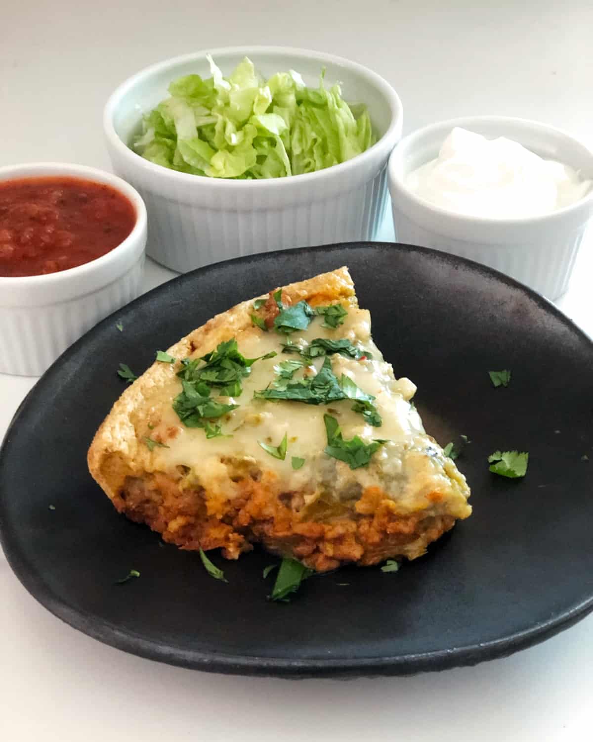 Slice of turkey taco pie on black ceramic plate with sides of salsa, shredded lettuce and sour cream in the background.