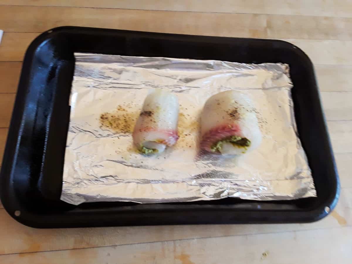 Tilapia fillets filled with basil pesto and rolled, sitting on foil-line baking pan.
