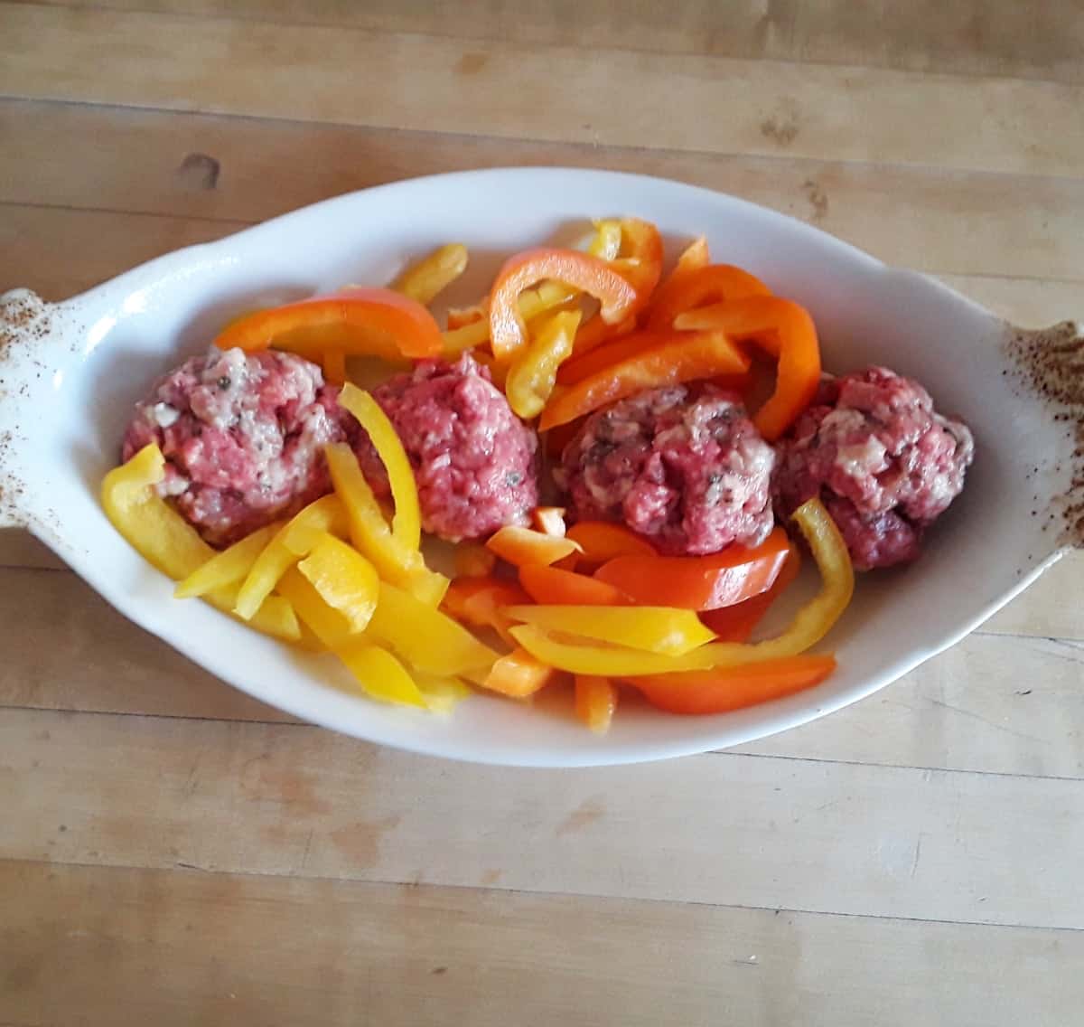 Uncooked meatballs with sliced peppers in small white baking dish.