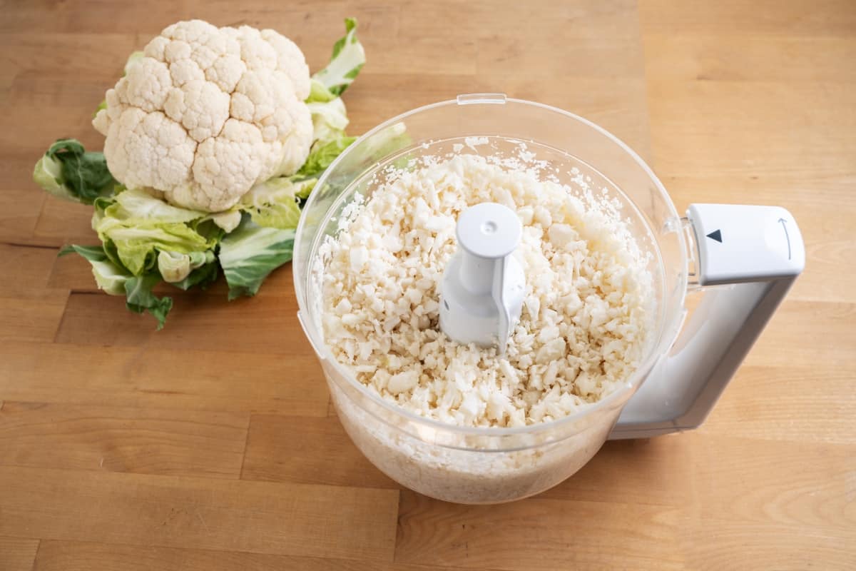 Food processor with riced cauliflower near head of fresh cauliflower.