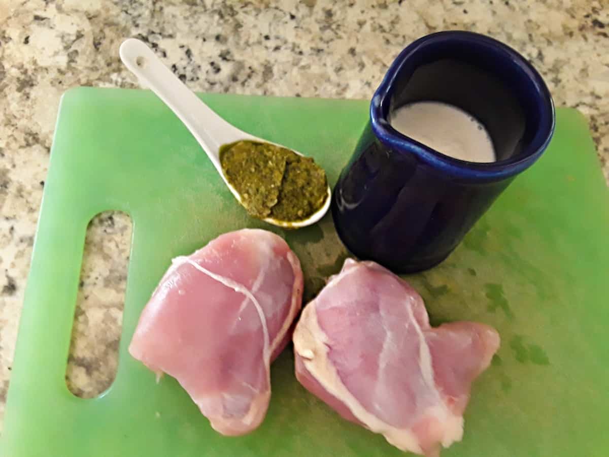 Chicken thighs, with pesto and coconut milk on green cutting board.