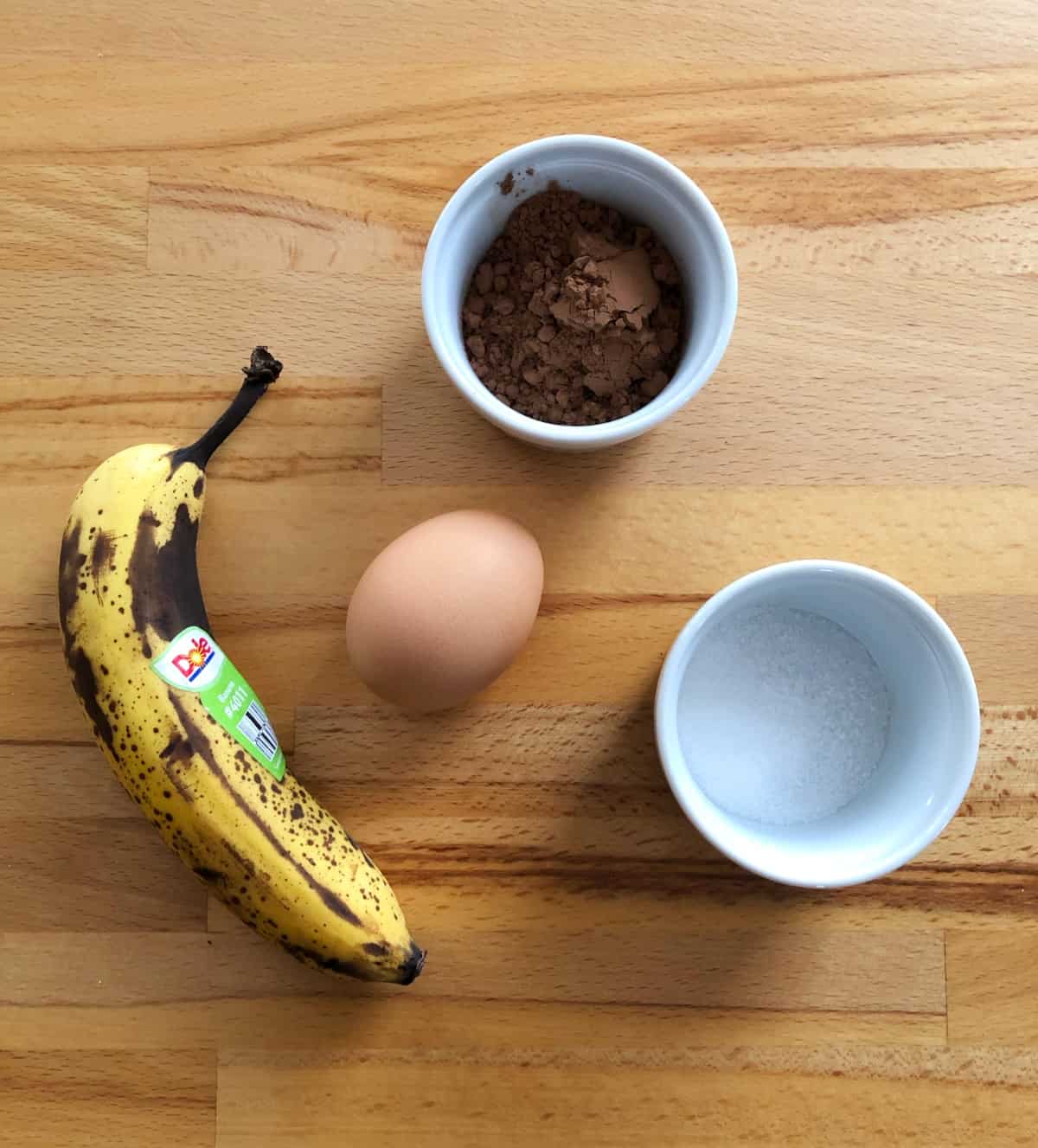 Over-ripe banana, cocoa powder, whole egg and sugar on wooden table.