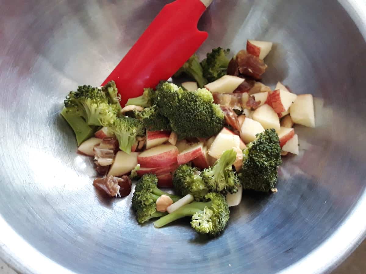Mixing to together chopped cooked broccoli with chopped apple in a stainless mixing bowl.