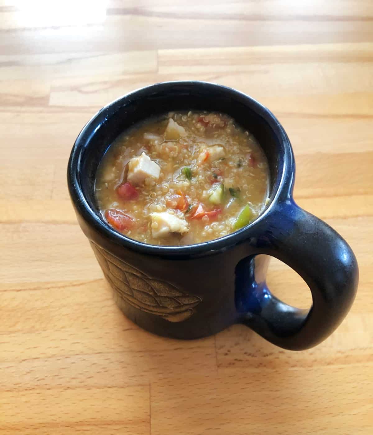 Chicken and quinoa soup in dark blue ceramic mug on wood table.