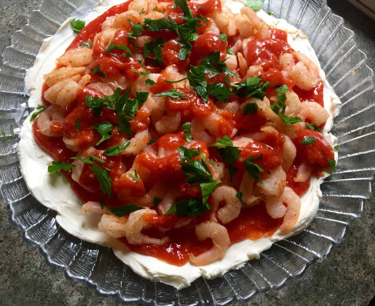 clear decorative glass plate spread with cream cheese and topped with small shrimp, cocktail sauce garnished with parsley