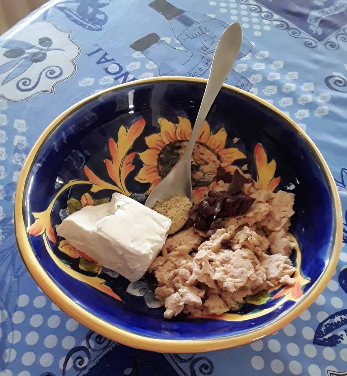 Cream cheese, tuna, olives and seasoning in blue mixing bowl with spoon.