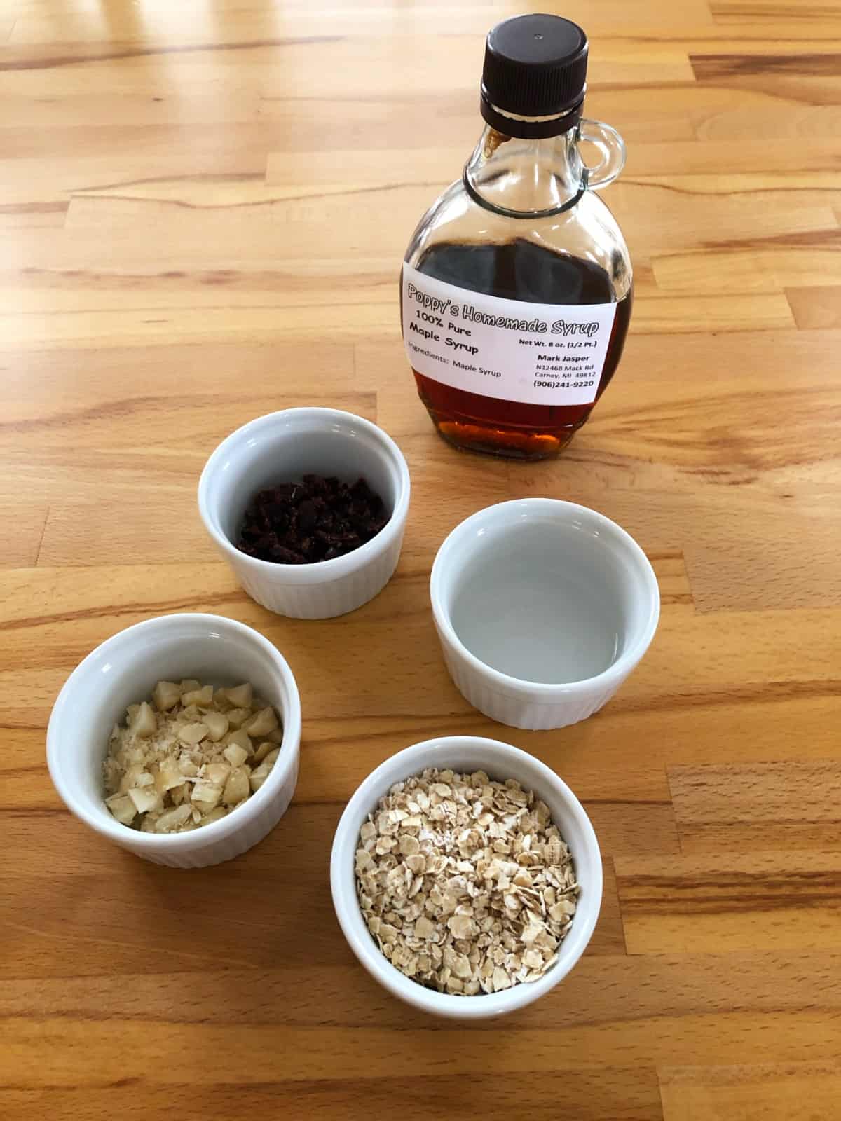 Ingredients for making granola, including maple syrup, oats, coconut oil, raisins and chopped macadamia nuts.