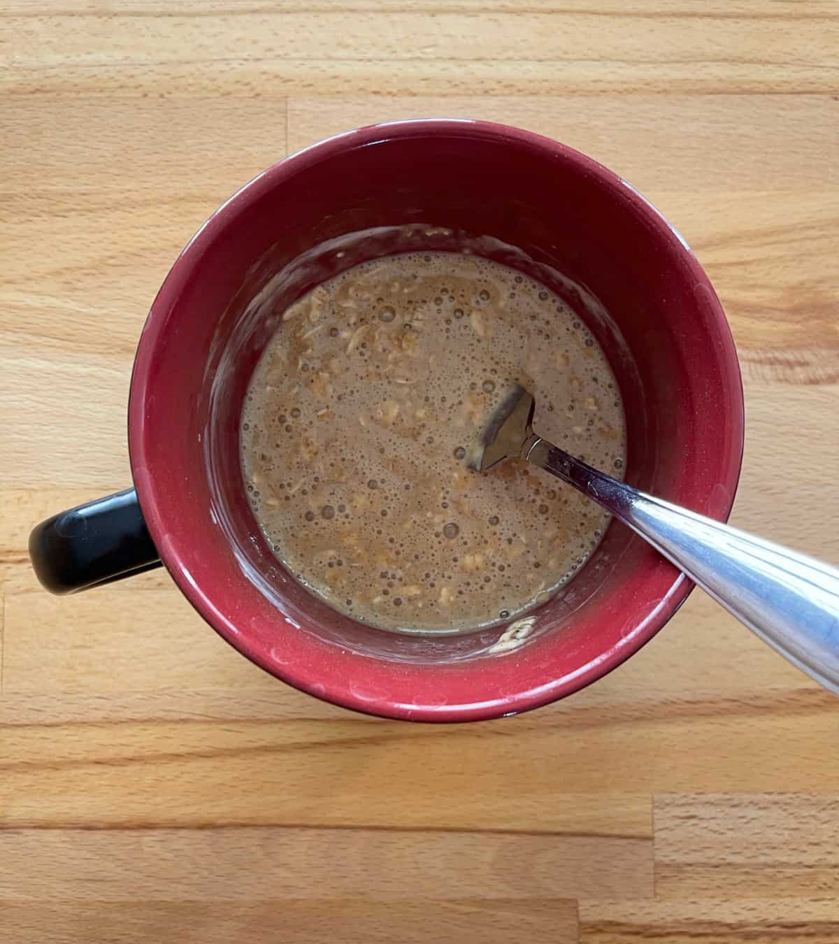 Mixing oatmeal raisin muffin ingredients in red mug with fork.