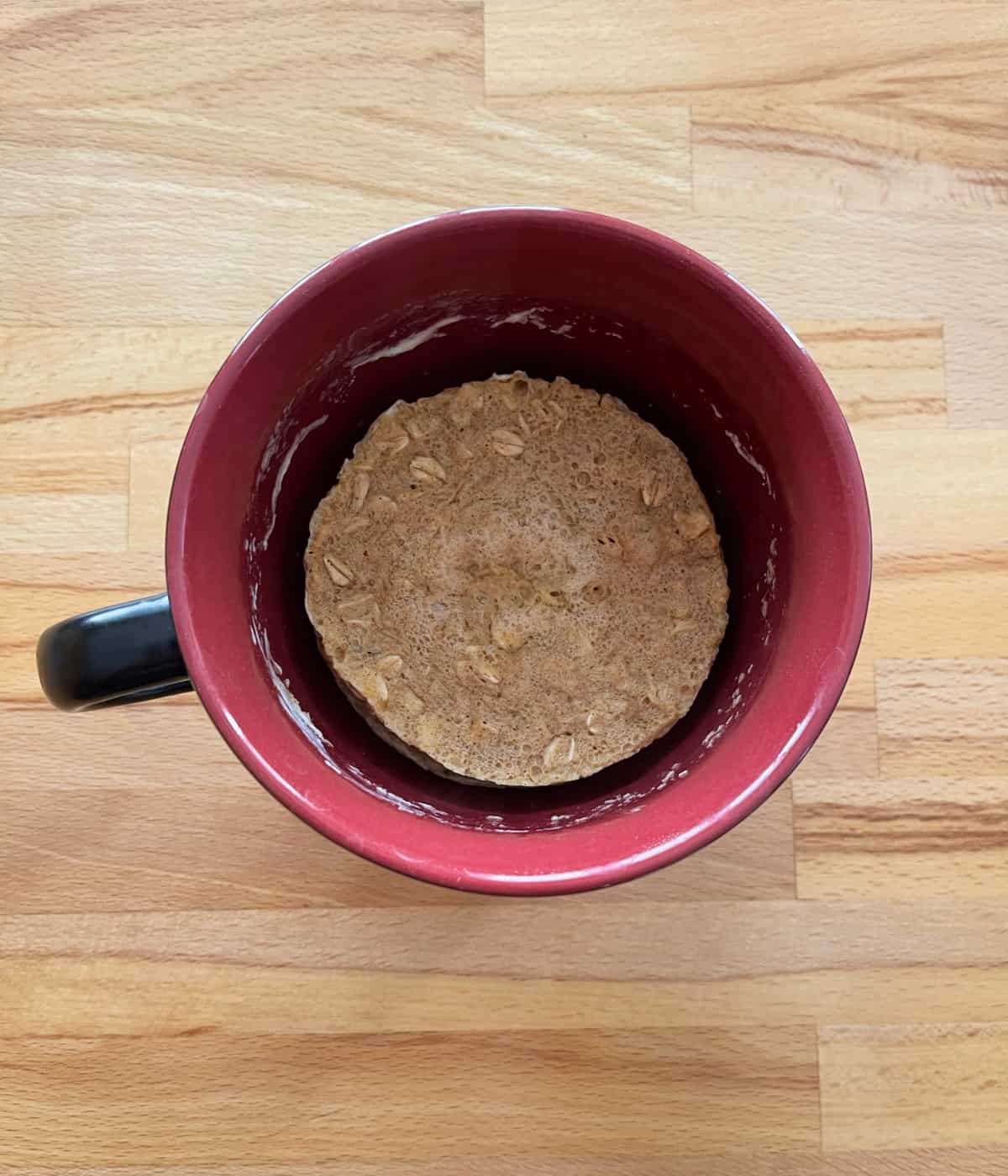 Oatmeal raisin muffin cooling in red mug on wooden table.
