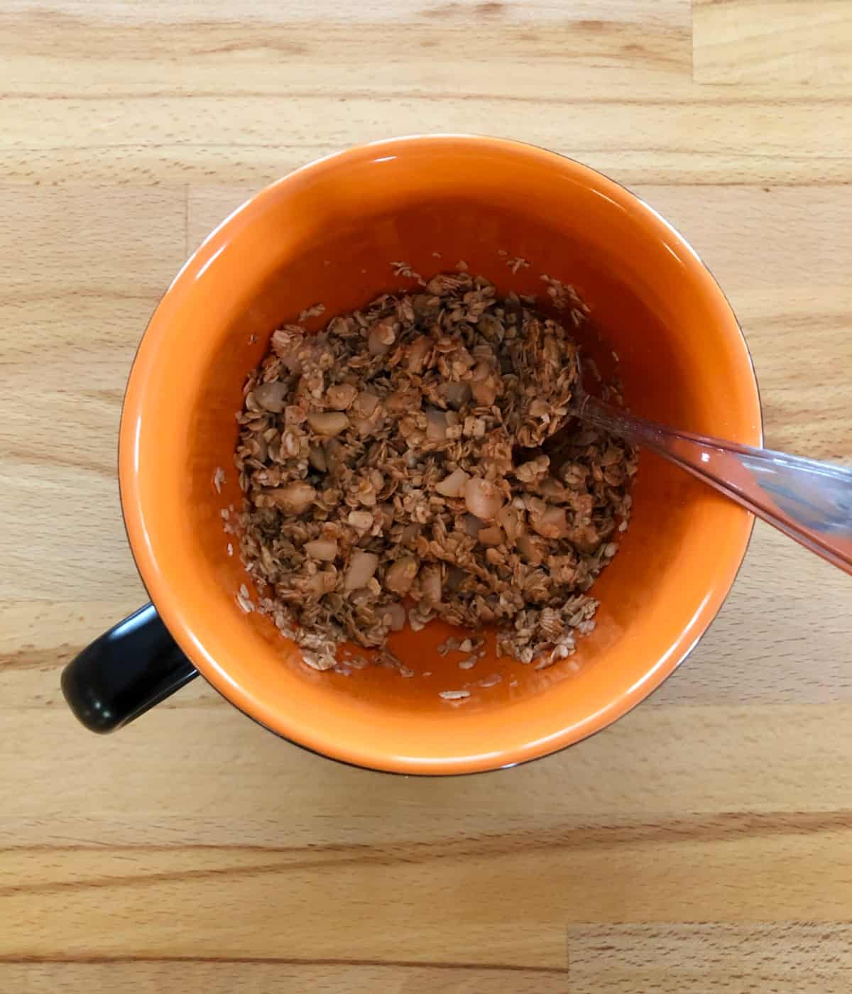 Mixing uncooked oats, maple syrup and coconut oil in orange mug with spoon.