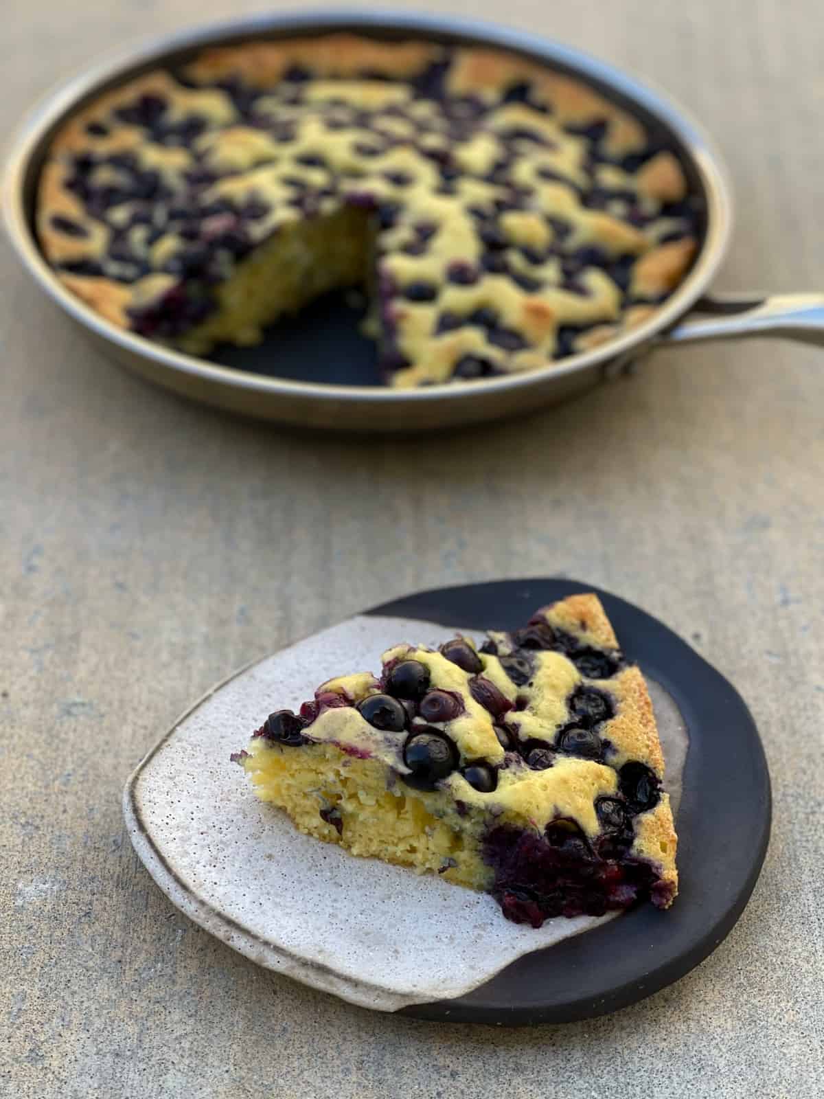 Wedge of blueberry oven pancake on ceramic plate with skillet in background.