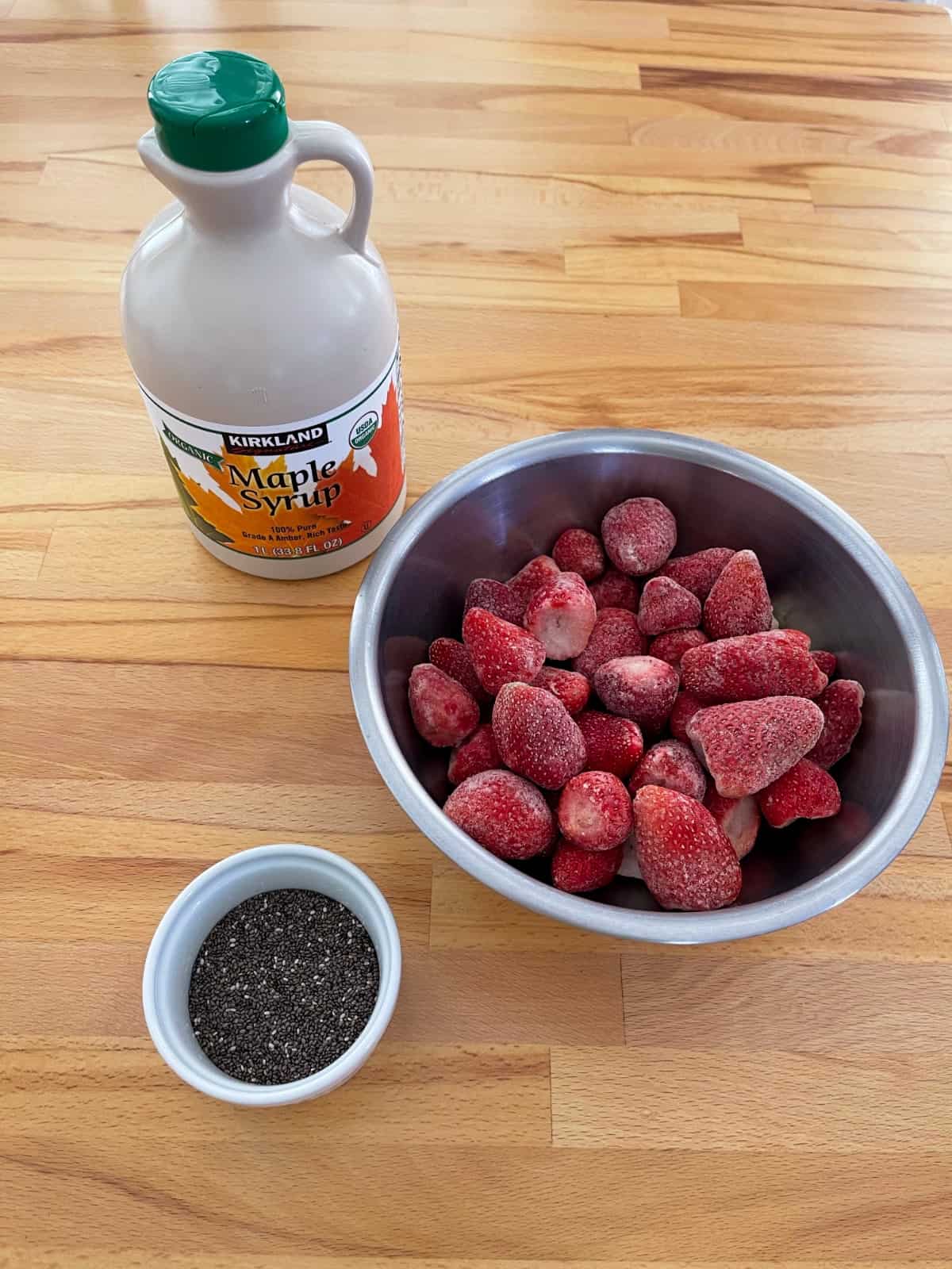 Pure maple syrup, frozen strawberries and chia seeds for making jam on wooden table.