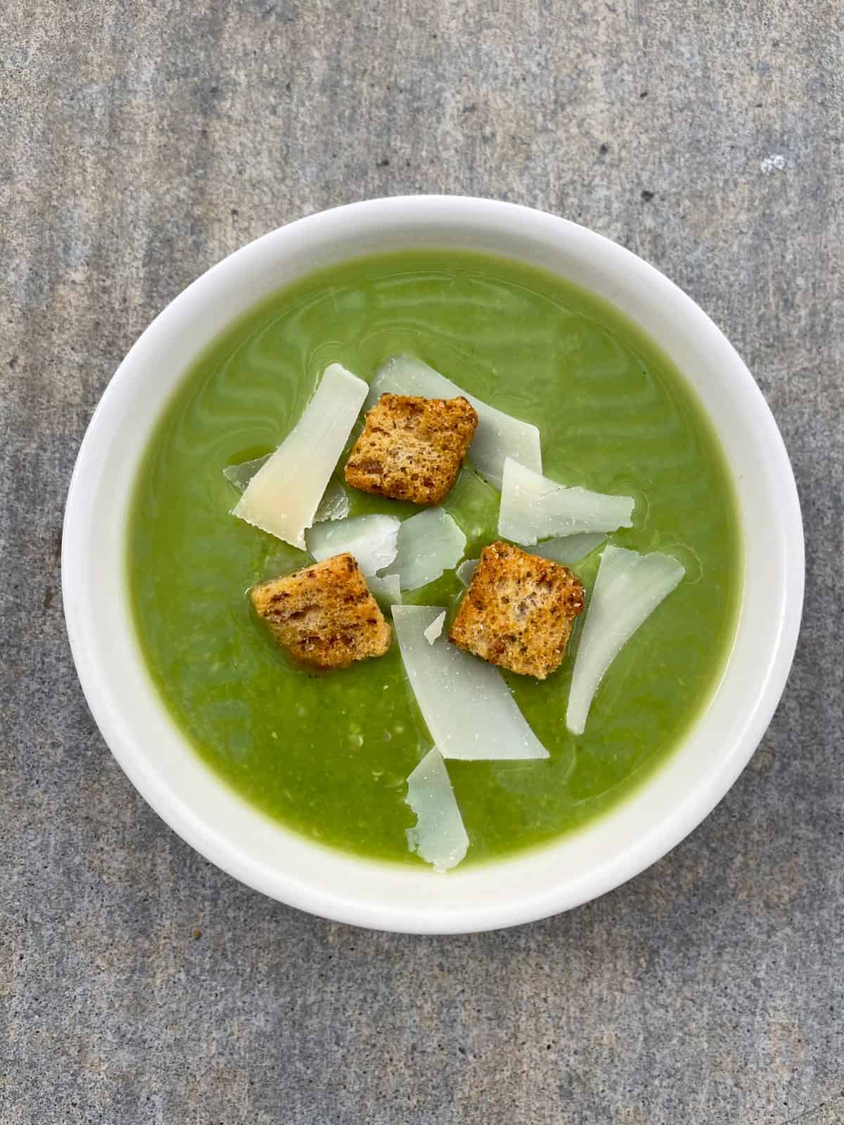 Quinoa pea soup in white bowl with croutons and shaved Parmesan.