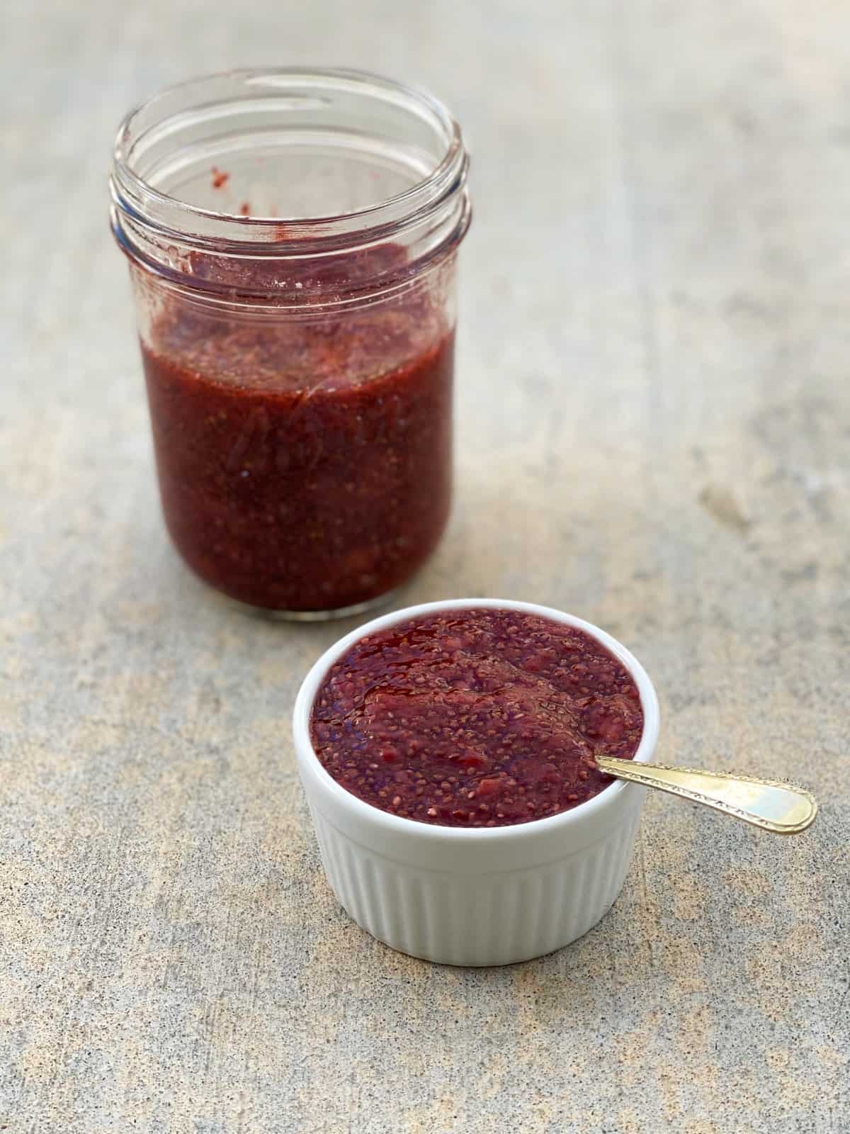 Strawberry chia jam in small white ramekin with spoon and large glass jar of jam in the background.