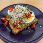 slice of toast topped with mashed avocado, sliced hard boiled egg, chopped cheery tomatoes, sprouts and crumbed bacon on a blue pottery plate